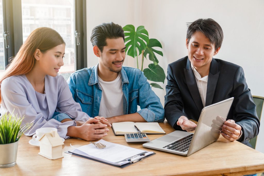 Dos clientes y un vendedor conversando el precio de venta unitario de un producto en una mesa con un ordenador y papeles.