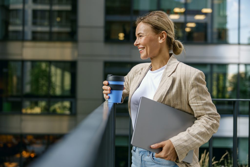 Mujer joven rubia con ordenador y café en la terraza de un edificio mirando al horizonte.