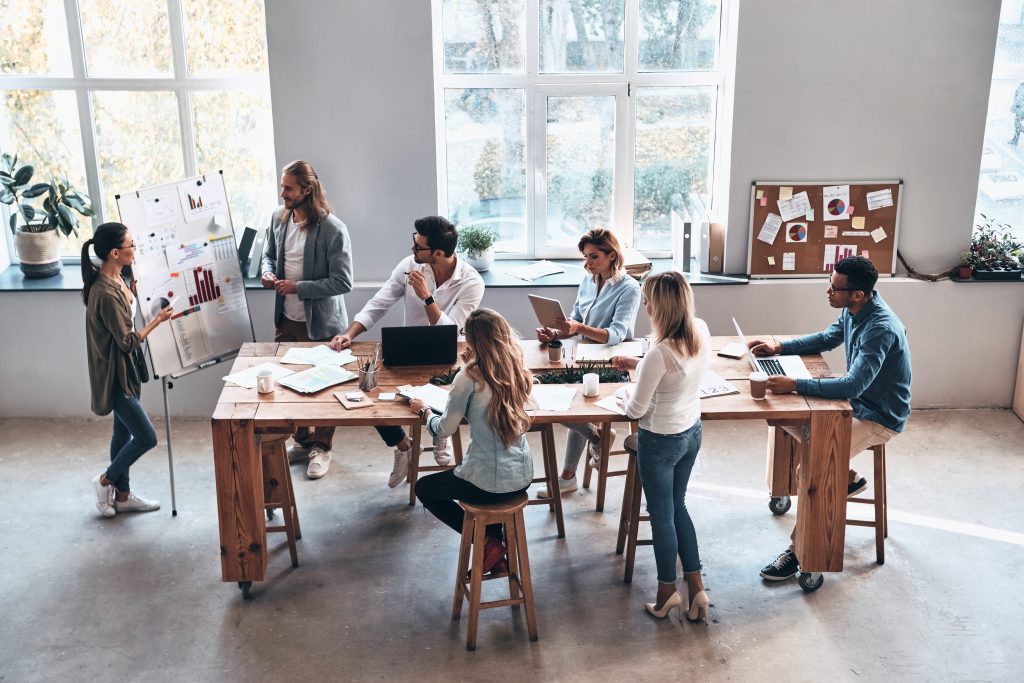 Grupo de compañeros de trabajo alrededor de una pizarra calculando el precio de venta de un producto en equipo alrededor de una mesa larga con ordenadores, tazas de café y sillas.