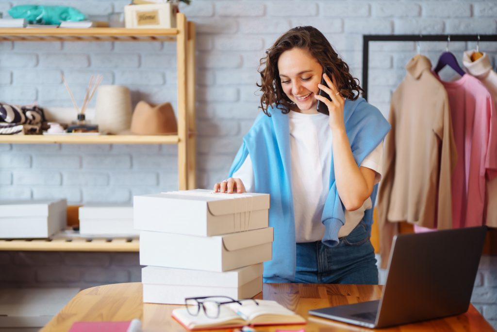 Dependienta de tienda dando un excelente servicio al cliente por teléfono mientras sostiene cajas sobre un escritorio para calcular el precio de venta